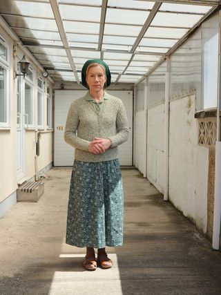 Juliet Stevenson as Dorothy Venn, wearing a cardigan, with a long skirt and a headscarf, standing outside one of the bungalows where the Barum Brethren live