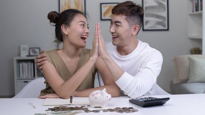 A young couple high-five while sitting at a table with a piggy bank and cash strewn before them.