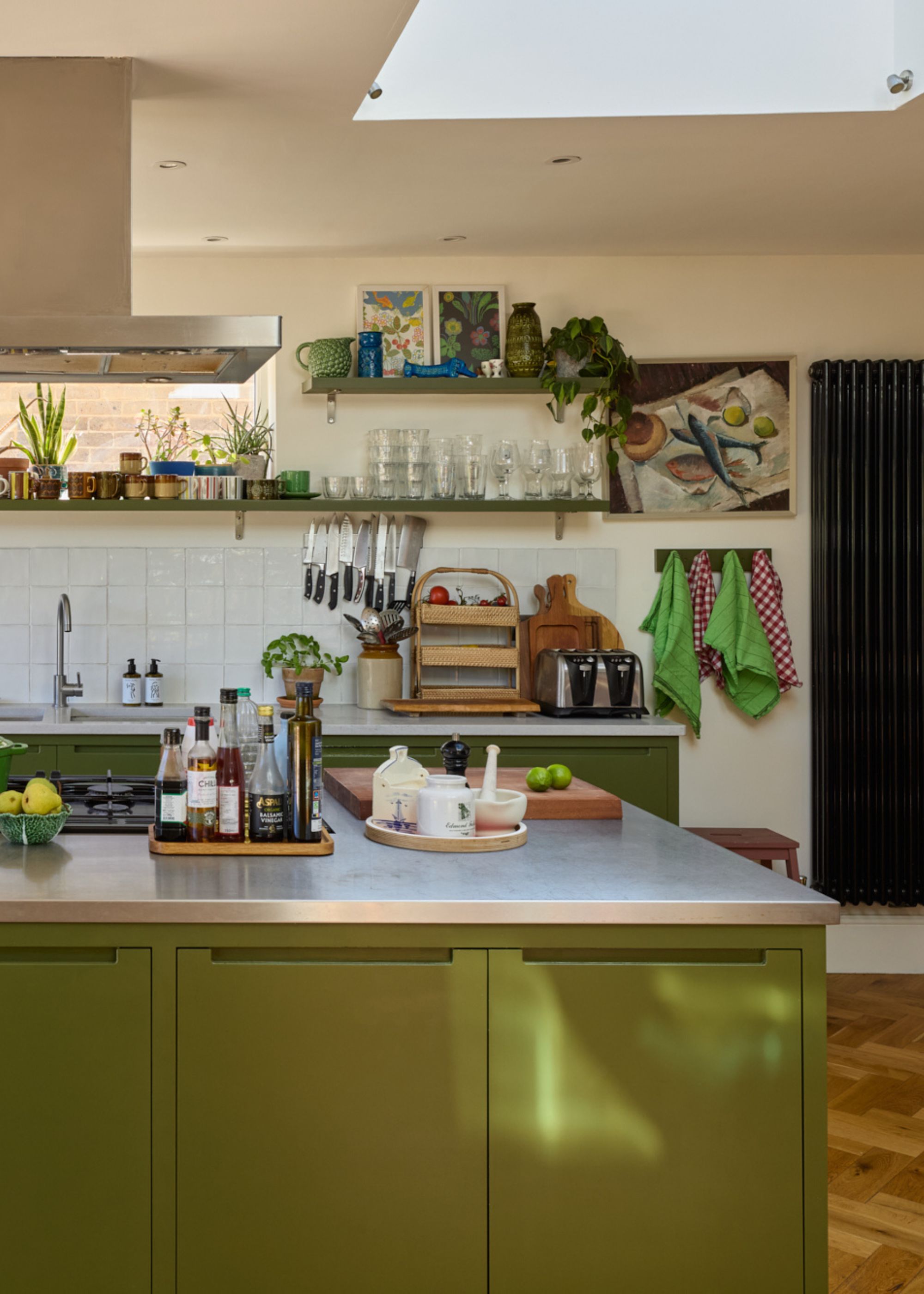 A modern kitchen with dark green cabinets on the kitchen island, stainless steel counters, white walls, and parquet wood flooring.