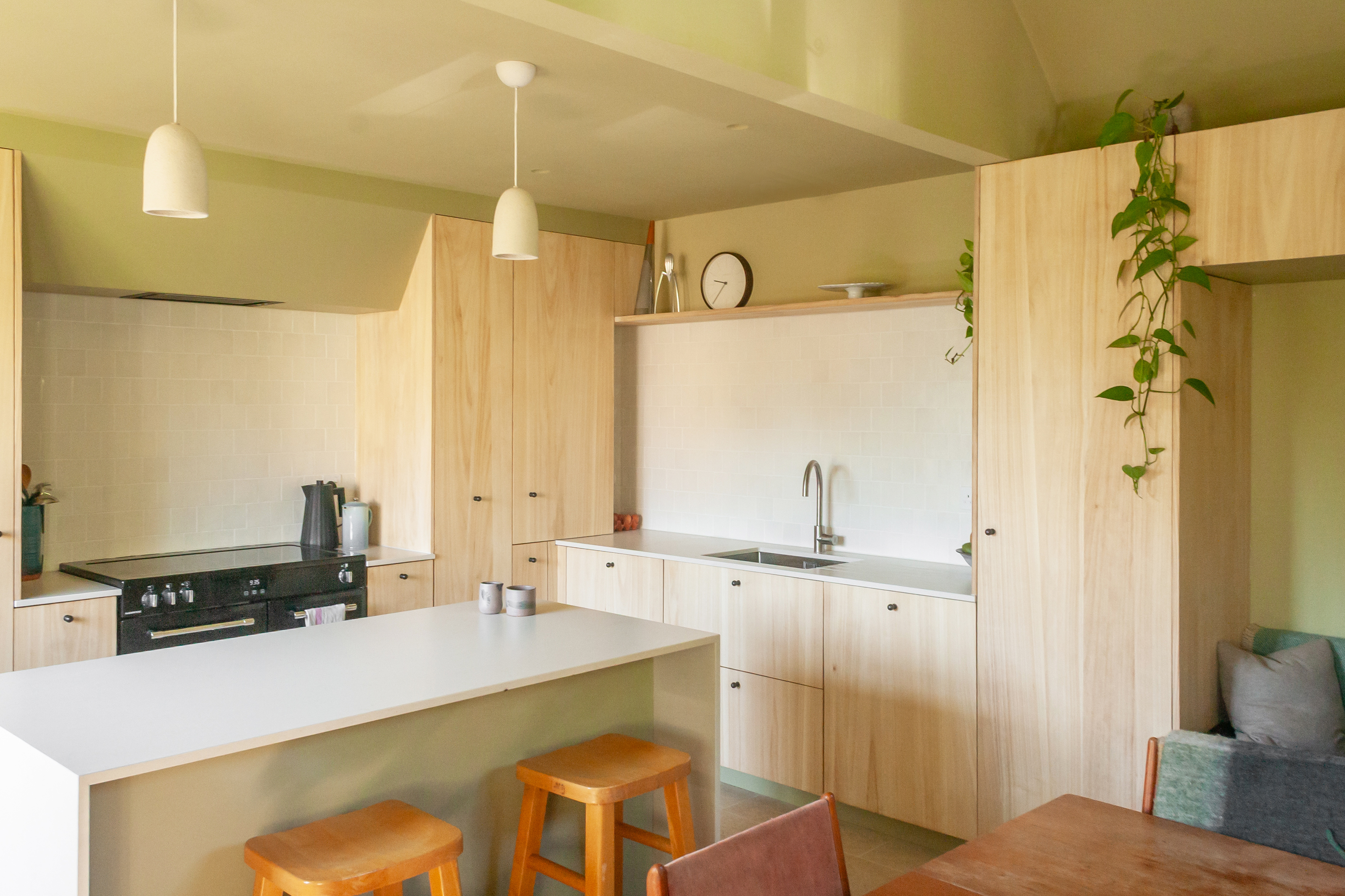A view of a modern, minimalistic kitchen with light wood cupboards, sage avocado green walls and ceilings, marble worktops, a kitchen island in the middle and dining area to the right, with a sitting nook in the right hand corner. 
