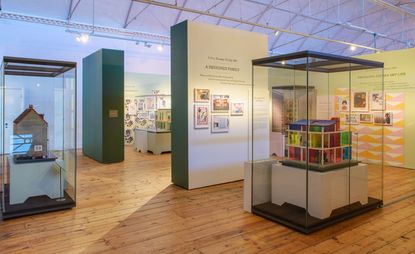 View of the 'Small Stories' show at the V&A Museum of Childhood featuring doll houses in glass display cases in a space with wood flooring and green and white walls with text and wall art