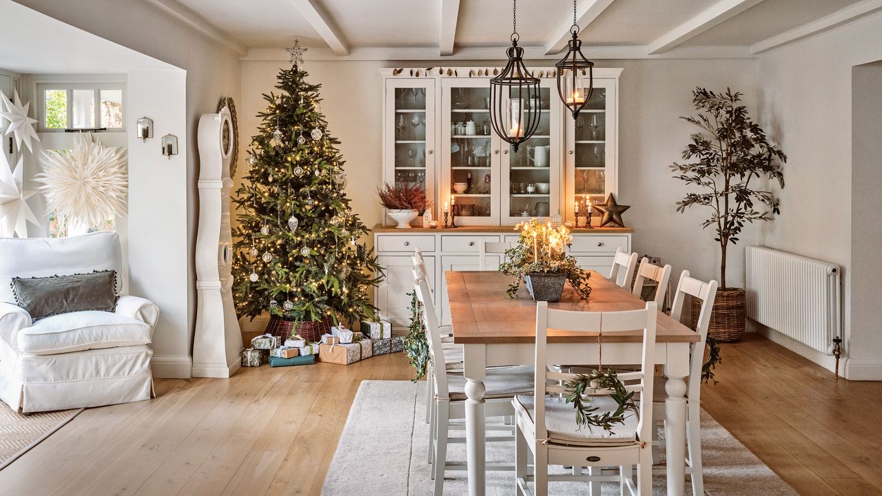 a dining room decorated for Christmas with sideboard and display case at one end of the table, a Christmas tree in the corner and white painted rustic dining furniture