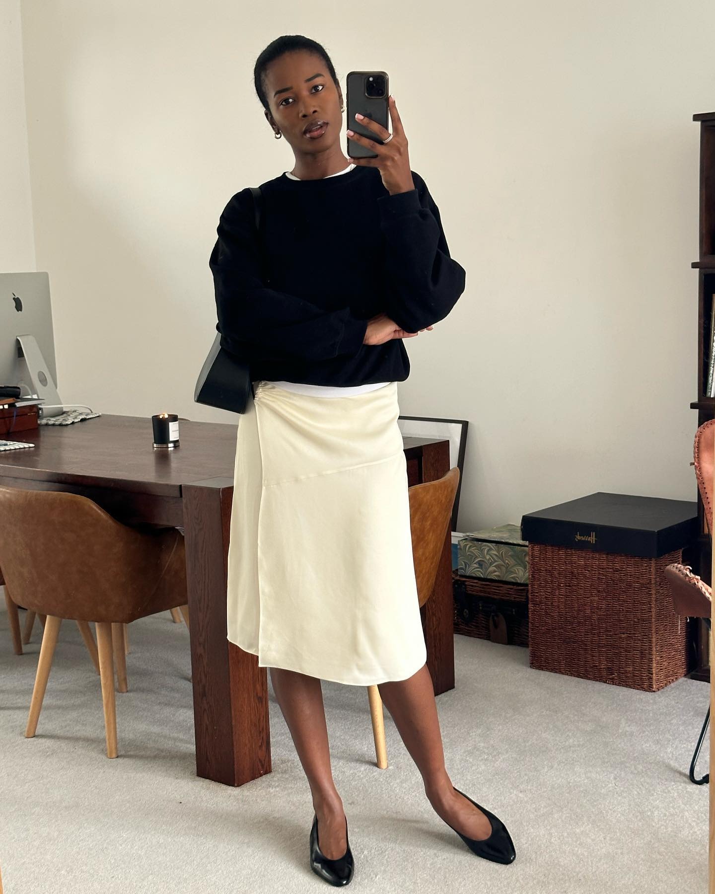 British style influencer Marilyn Nwawulor-Kazemaks poses in her chic London home wearing a black crewneck sweater, a beige wrap skirt, and almond-toe kitten heels