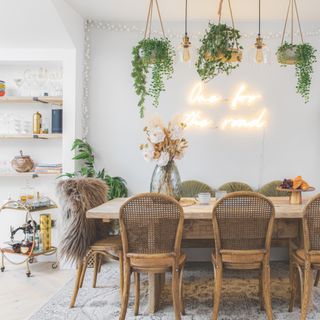 small dining room area next to kitchen, large dining table with chairs one side, bench the other, plants hanging above table with glass shade pendants, neon sign on wall, rug, sheepskins, drinks trolley, open shelving