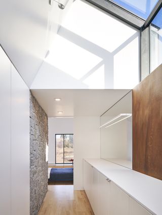 Interior view of a space at Smith House featuring white walls with stone and wood sections, a skylight, wood flooring, a wall mirror, spotlights and a white storage unit. There is a partial view of a bedroom