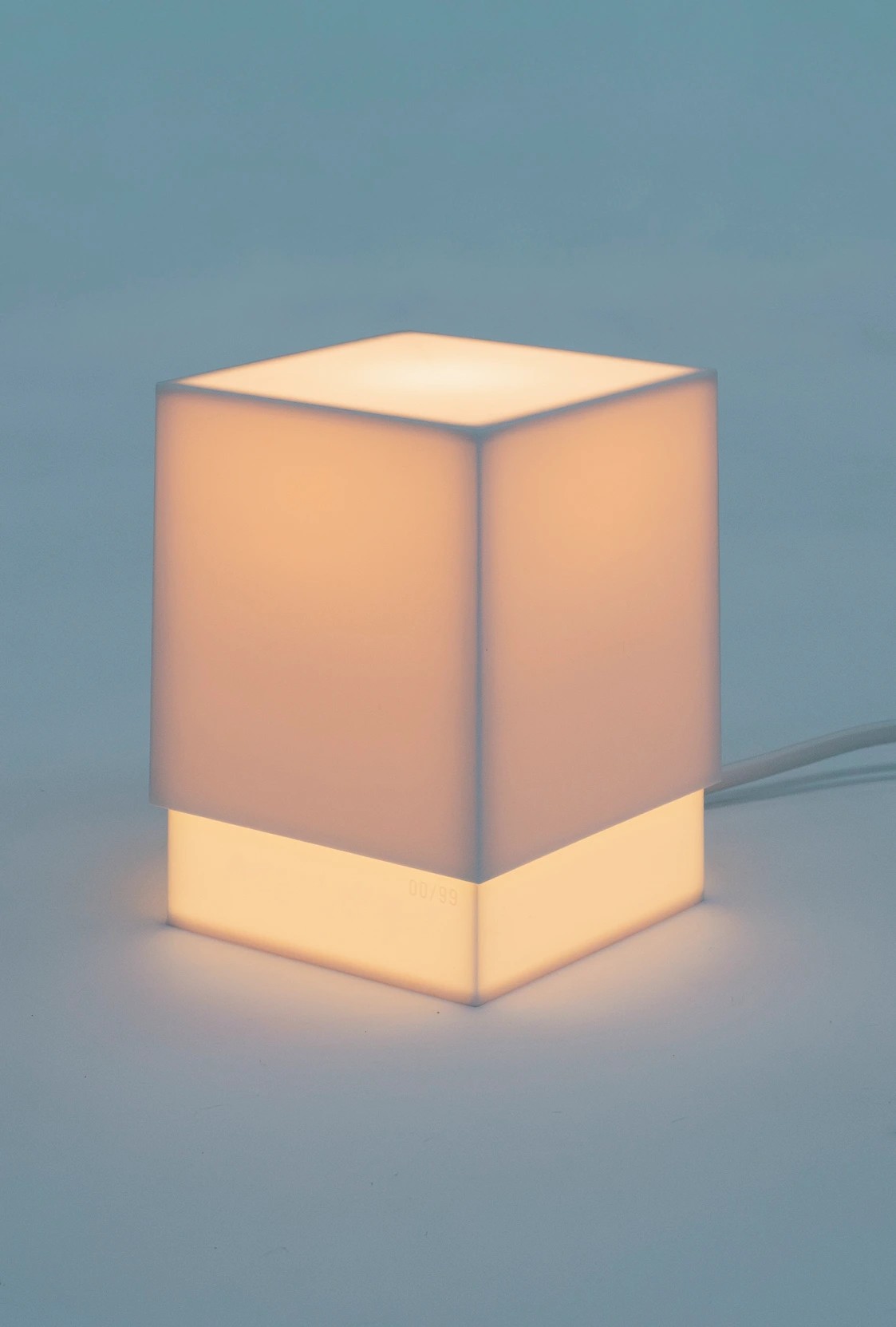 A square, box-like table lamp set against a pale blue background glows with pale pink light.