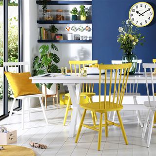 A blue, white and yellow dining area with a table and wooden chairs and wall clock