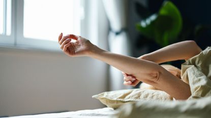 Woman with arm raised over bed in the morning light, body under the duvet, representing what is edging and its benefits