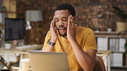 A man massages his temples as if he has a headache while looking at his laptop.