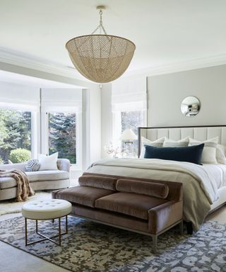 Modern master bedroom with gray painted walls, large bay window, daybed at the foot of bed, large golden pendant light hanging over bed, gray carpet with large patterned rug, rounded mirror on wall behind bed