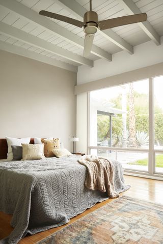 A large bedroom with high white wooden ceilings and a ceiling fan, big windows, and lots of cushions on the bed.