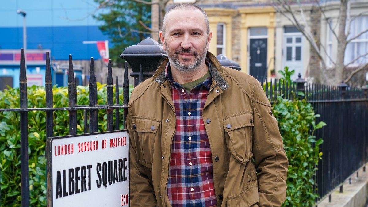 Alex Walkinshaw as Ross in EastEnders leaning against the Albert Square sign in a check shirt and brown jacket