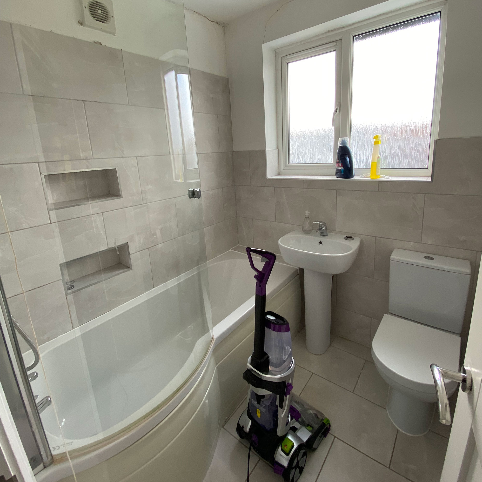 Grey bathroom with white fixtures, glass shower screen and vacuum on floor