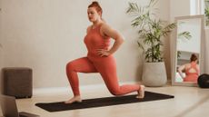 A woman in a sports vest and leggings performs a lunge at home on an exercise mat. Her right foot is planted on the mat, right knee bent, while her left leg is behind. Her hands are on her hips. Behind her we see a floor length mirror and potted plant.