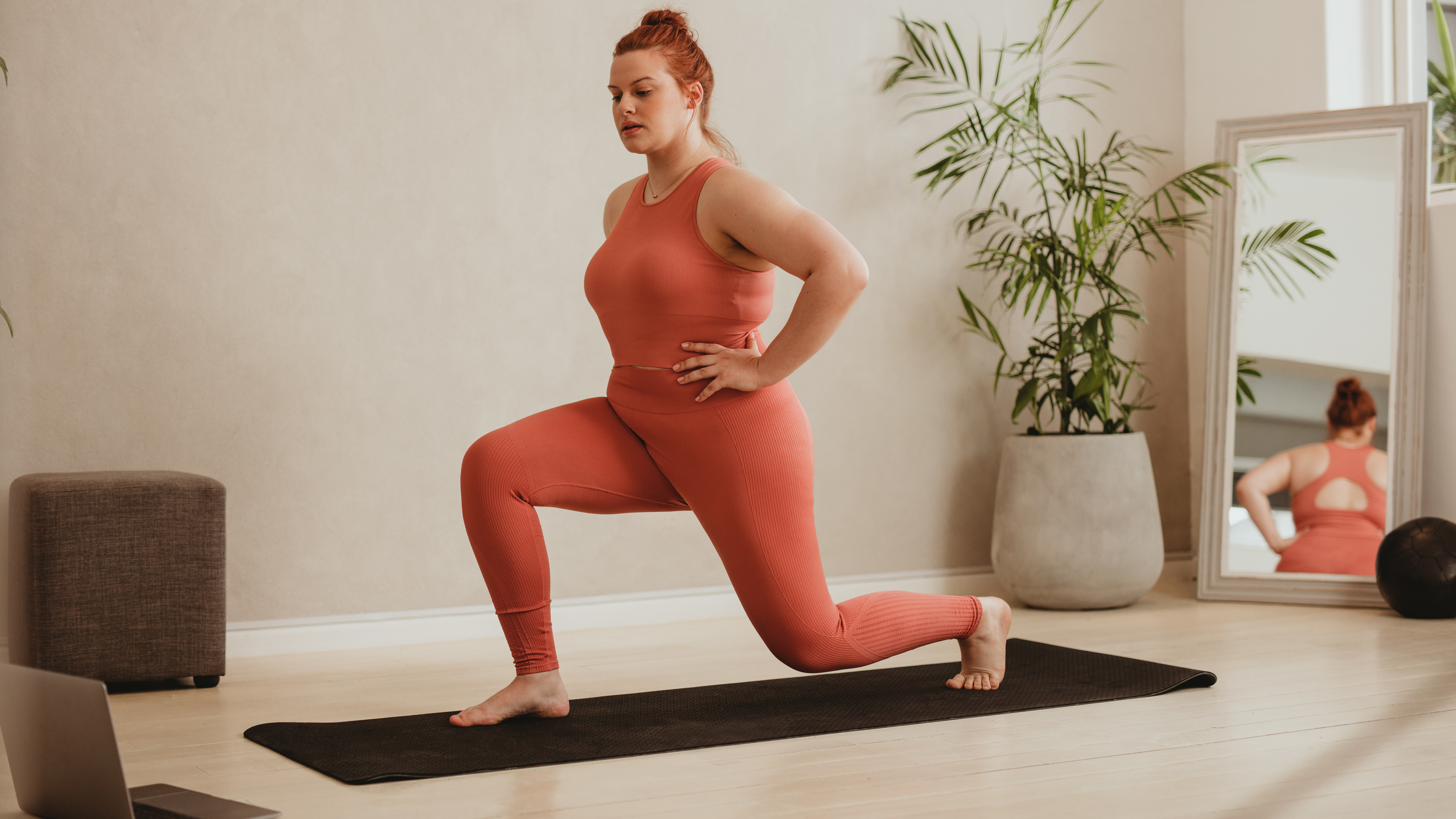 A woman in a sports vest and leggings performs a lunge at home on an exercise mat. Her right foot is planted on the mat, right knee bent, while her left leg is behind. Her hands are on her hips. Behind her we see a floor length mirror and potted plant.