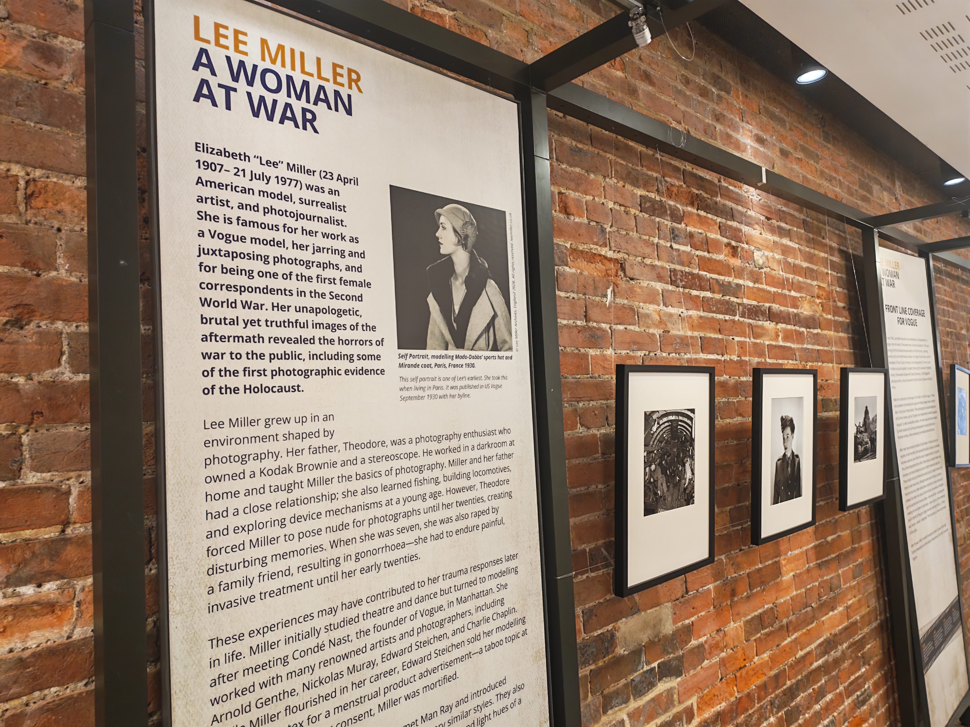 The exhibition's introductory panel, &amp;quot;Lee Miller: A woman at war,&amp;quot; presents a biographical overview beside a self-portrait of Miller in Paris, with framed wartime photographs receding along the brick wall behind