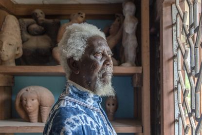 Photograph of Demas Nwoko working in his studio, dressed in a long blue and white pattern over shirt, wood framed and pattern window, grey wall, wooden shelving with pottery sculptures in the backdrop - the architect won the Golden Lion for Lifetime Achievement 2023