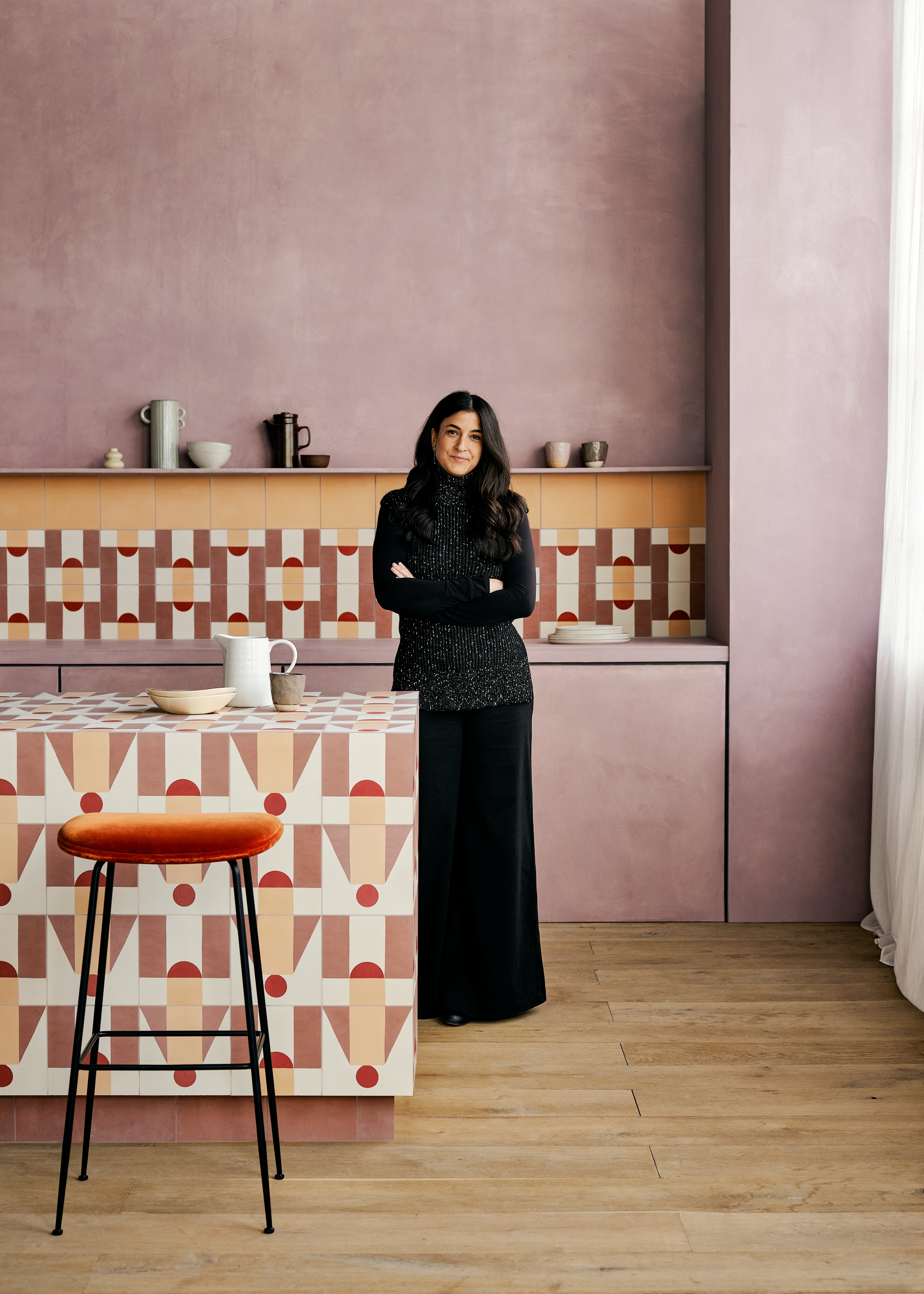 A woman dressed in a smart long skirt and a heavy black and white jumper stands in a rose-tinted kitchen with wooden floors, tiled surfaces in tones of argilla, pale nude, and white, high velvety orange stools, and crafty ceramics.
