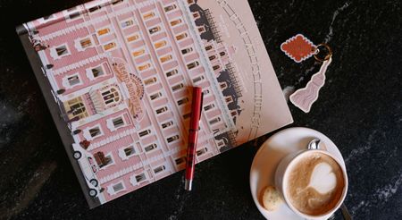 A pink book depicting a palatial hotel's facade, topped by a red pen and sitting next to a capuccino mug, is shot from above.
