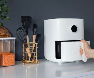 A white air fryer on a white countertop with kitchen utensils and red lentils beside it