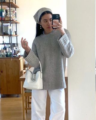 Fashion influencer Sasha Mei poses for a mirror selfie wearing a bandana, ball jewelry, a gray crewneck sweater, a white bag, and white jeans.