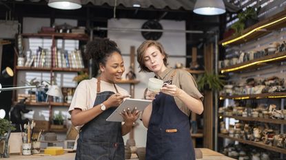 Two women wearing aprons consult a tablet in a craft store.