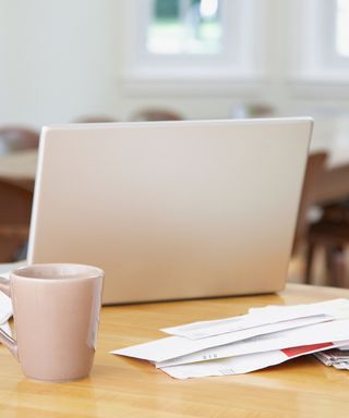 A wooden table with a silver laptop, a light pink mug, and a pile of white envelopes and paper on it, with a white wall and windows in the background