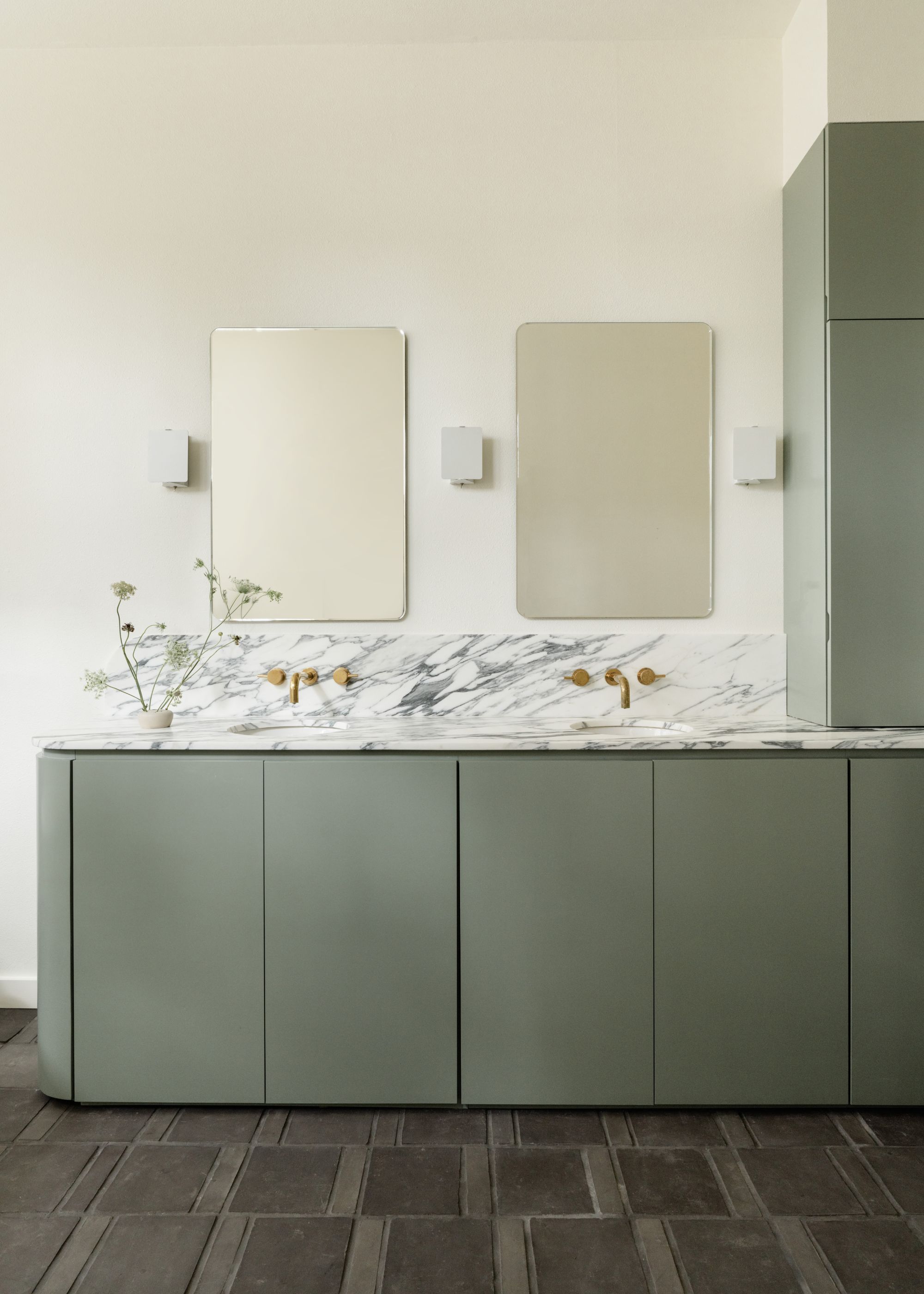A minimalist, modern bathroom with a muted green vanity, off-white walls, double sinks with marble countertops and two rectangular mirrors on the wall.