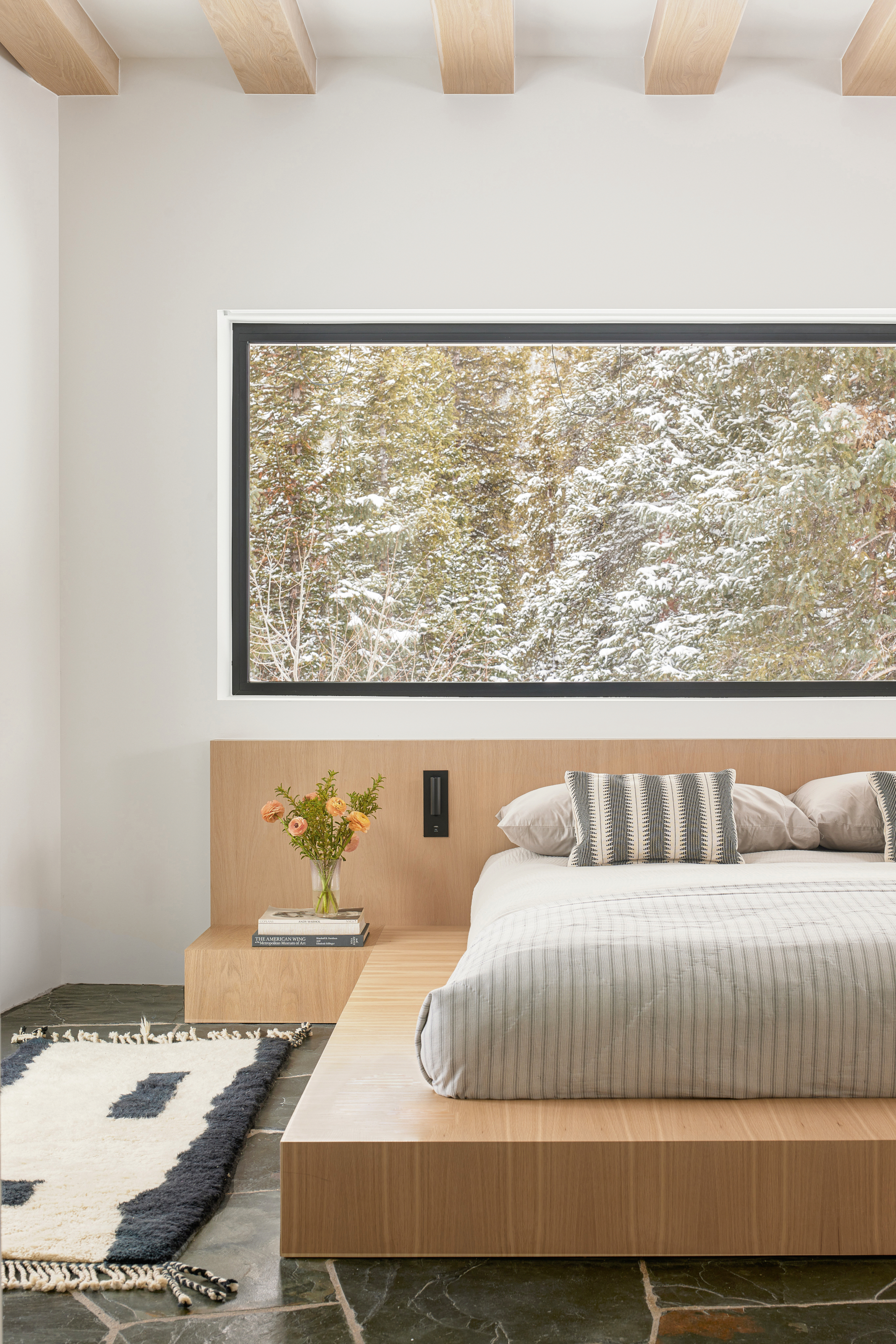 A bedroom with built-in platform bed in white oak, stone floor, black and white kilim rug and picture window overlooking snowy trees