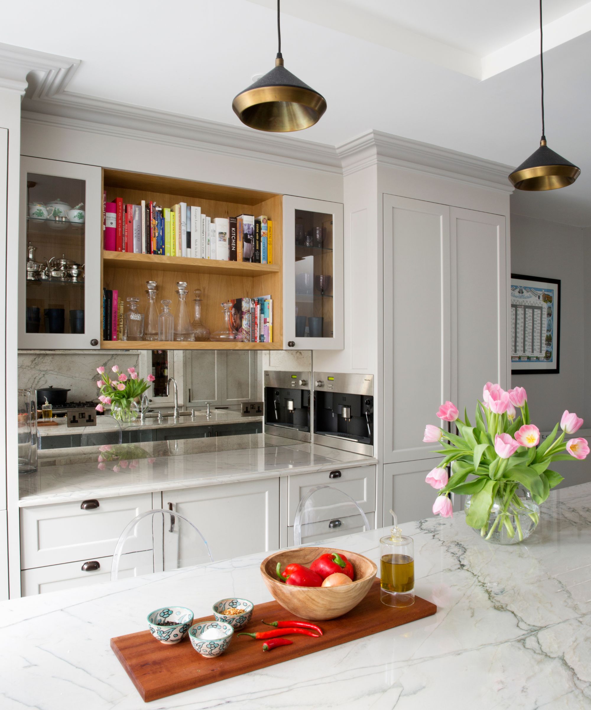 A white kitchen with marble countertops, white cabinets, wooden shelves, a cutting board and pink tulip flowers in a glass vase.