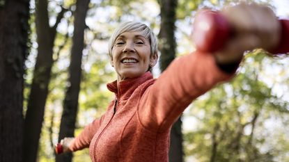 An older woman smiles as she lifts weights in a wooded setting.
