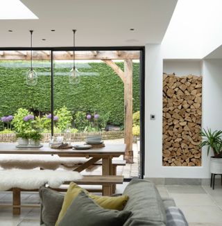 logs stacked in an indoor log store next to a wooden dining table and glass doors with garden views