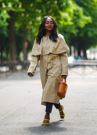 Carrole Sagba @linaose wears gold earrings, a long beige coat with a cloak collar, blue denim jeans pants, an orange shiny leather handbag with a gold shark teeth buckle, yellow shiny leather strapped block heel sandals, on May 02, 2021 in Paris, France