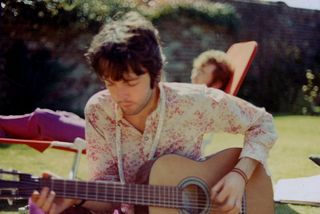 Paul McCartney from the Beatles plays an acoustic guitar while John Lennon (1940-1980) sunbathes behind in London, summer 1967.