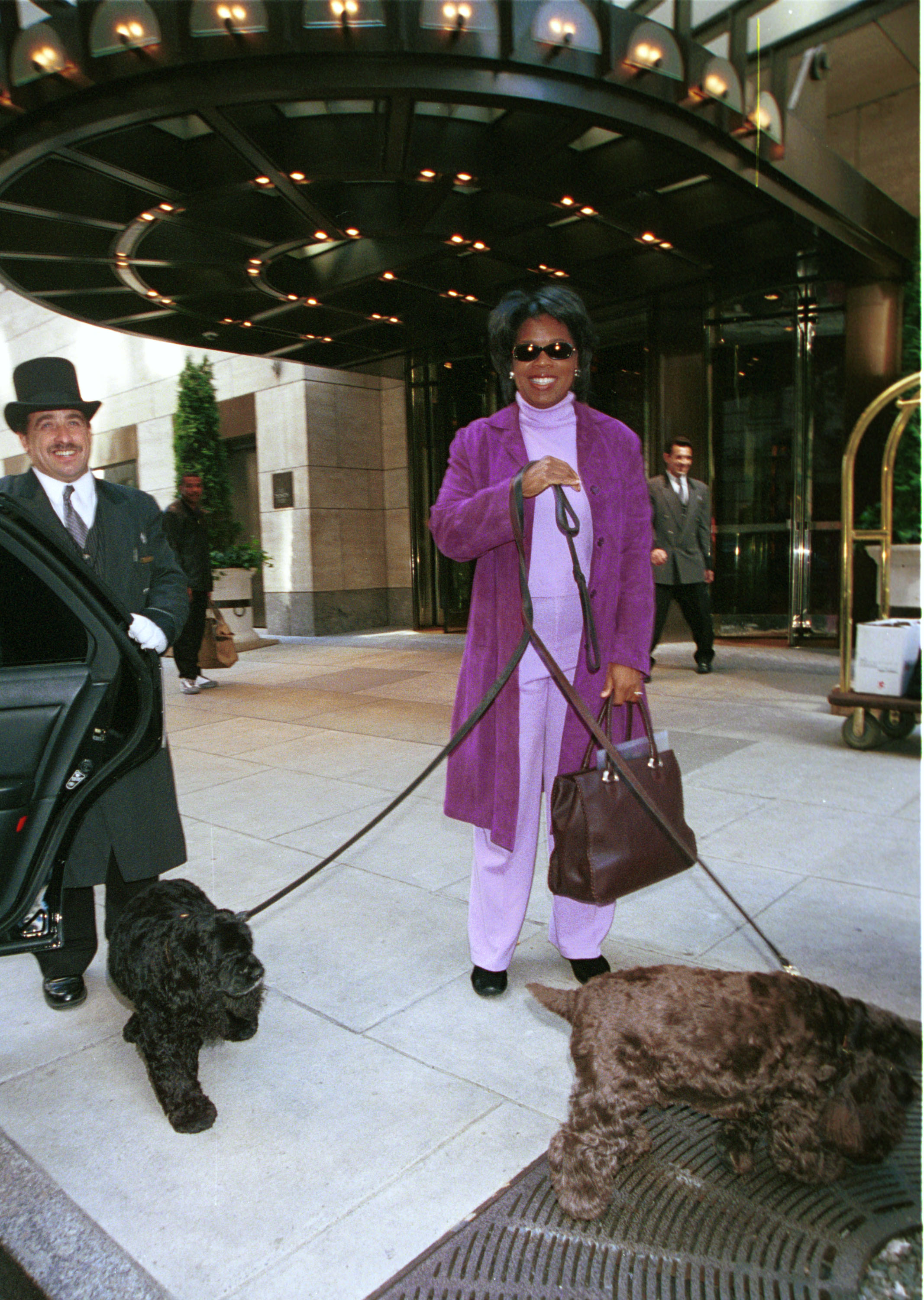 A woman dressed in lilac and purple standing outside a hotel entrance, smiling as she holds two brown Cocker spaniels on leads.