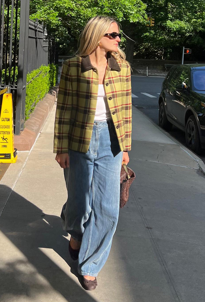 A woman walking down the sidewalk in New York City wearing a yellow and brown plaid jacket over a white tank top, light-wash barrel-leg jeans, brown ballet flats, and a brown handbag.