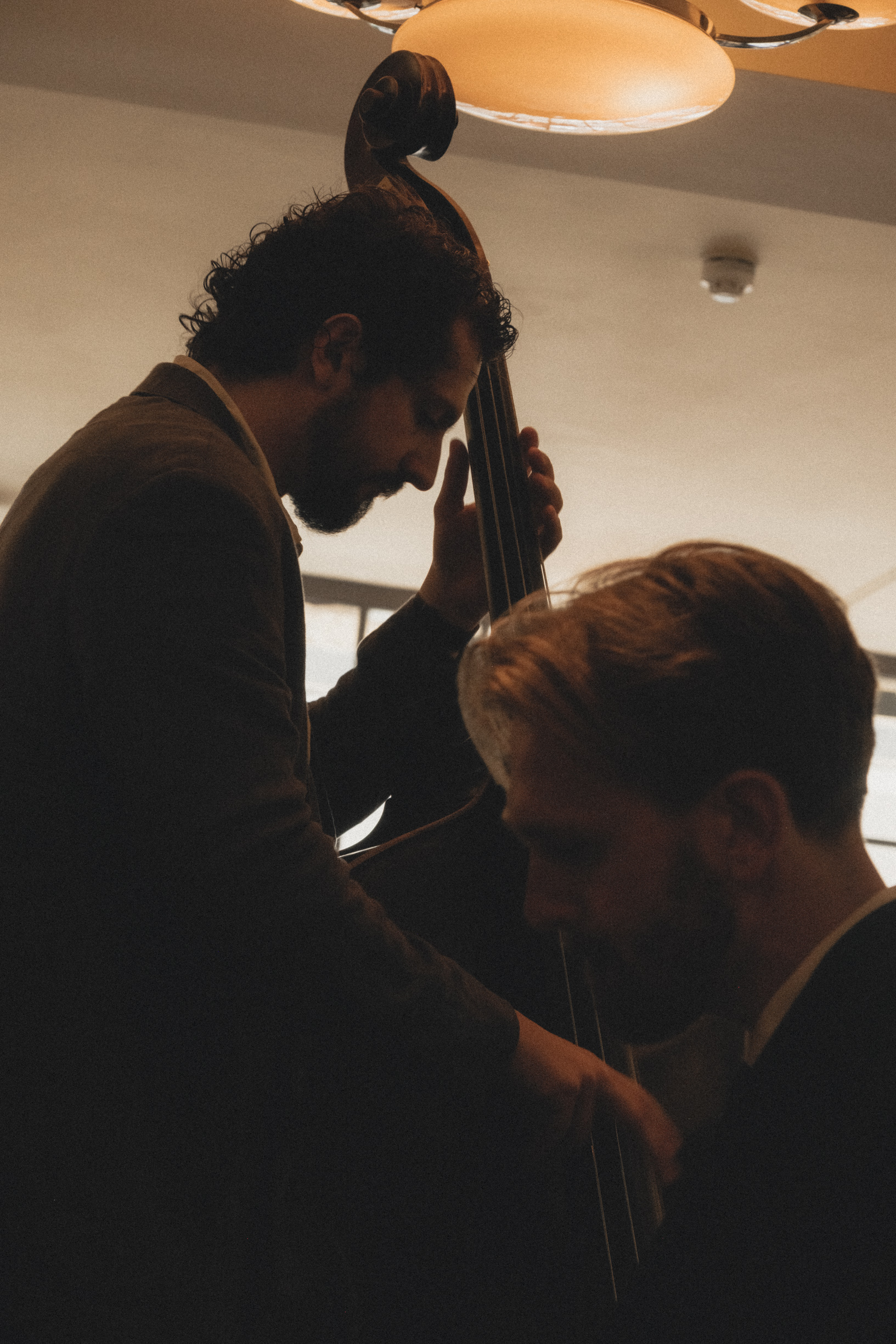 A jazz ensamble made up of two men dressed in suits playing the cello in a dimly lit restaurant with amber-hued pendant lights.