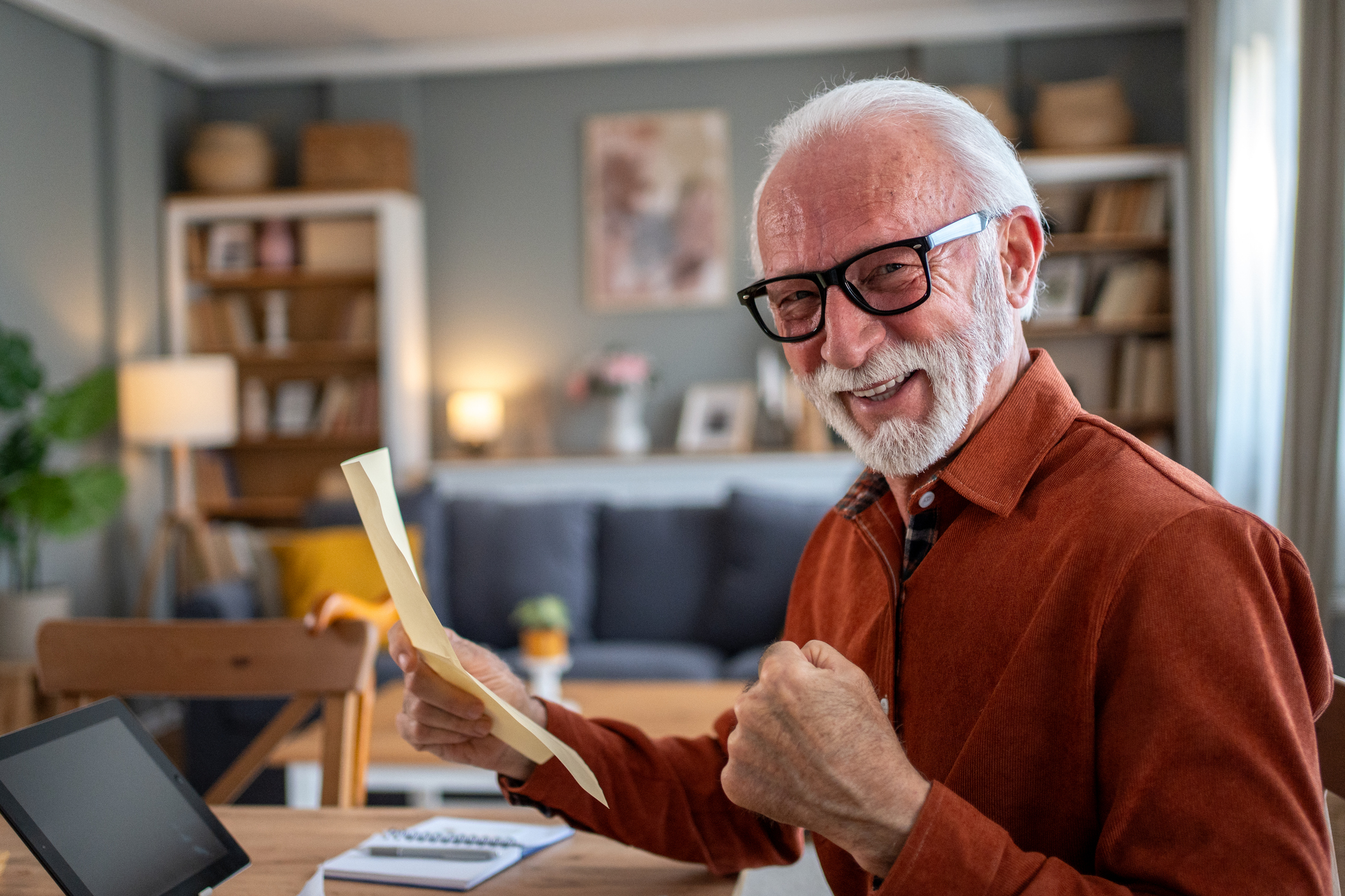 Cheerful senior man sitting at home desk, holding a document while expressing joy and success with a triumphant fist pump, radiating happiness and excitement in his cozy home office