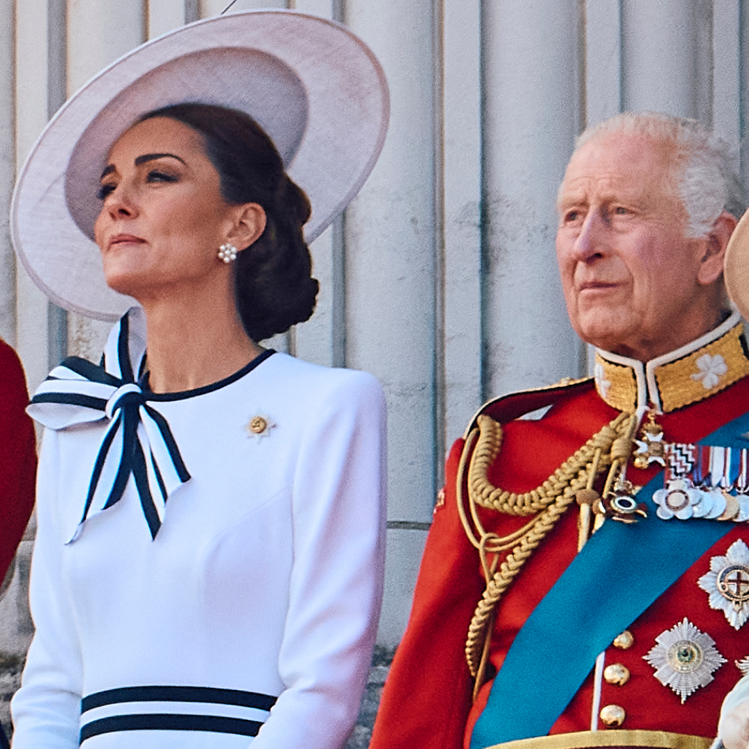 Prince William and King Charles wear red military uniform and Kate Middleton wears a white dress with navy accents and a matching hat on the Buckingham Palace balcony