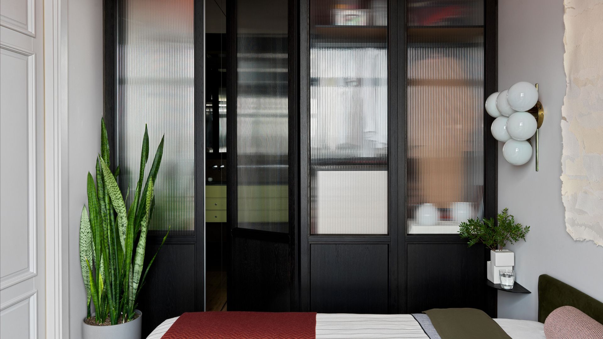 A bedroom with a glass closet featuring ribbed glass by a washroom. Also in the room is a potted snake plant, a floating side table with a white vase of stems below a bubbled wall sconce, beside a hanging painting