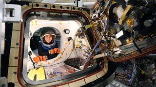 an astronaut with a helmet visible in the round doorway of a spacecraft hatch, whose door is open showing gears and mechanics