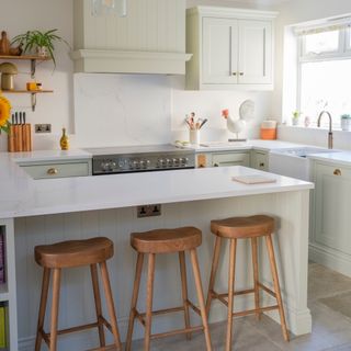 pale green and white kitchen with wood bar stools, stone floor, white worktops, wall units