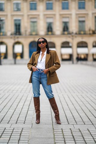 A woman wears a tan blazer, white button-down shirt, jeans, and brown knee-high boots.