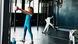 Woman holding a kettlebell in two hands in the middle of a kettlebell swing, smiling at the gym