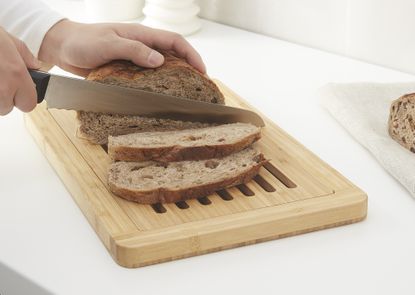 A close up of a cutting board on a kitchen countertop with a person cutting bread with a knife