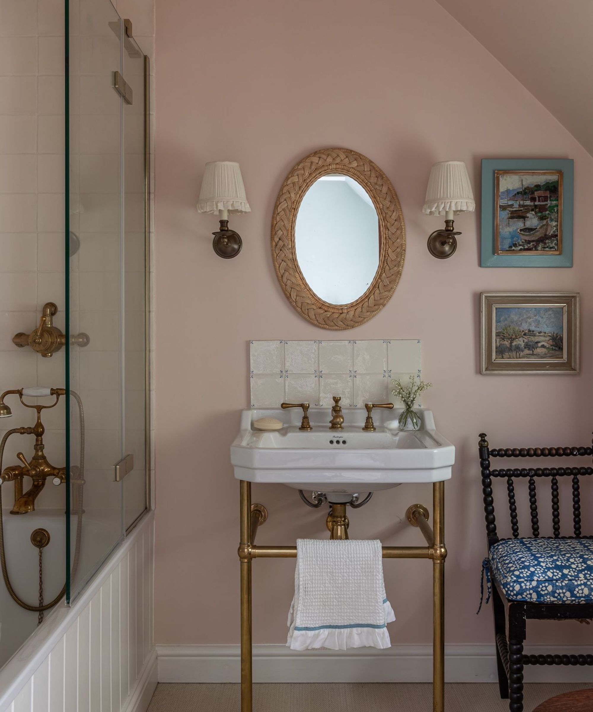 Pale pink bathroom with brass sink, rattan mirror, sconces, vintage paintings and a wood panelled tub