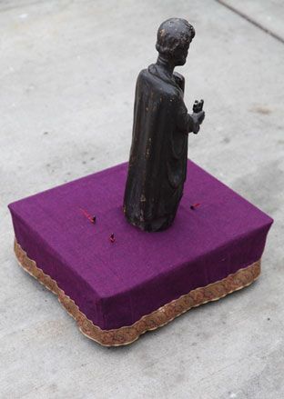 Grey paved floor, bronze caped male sculpture, set on a plinth of purple cloth with multicoloured lace trim