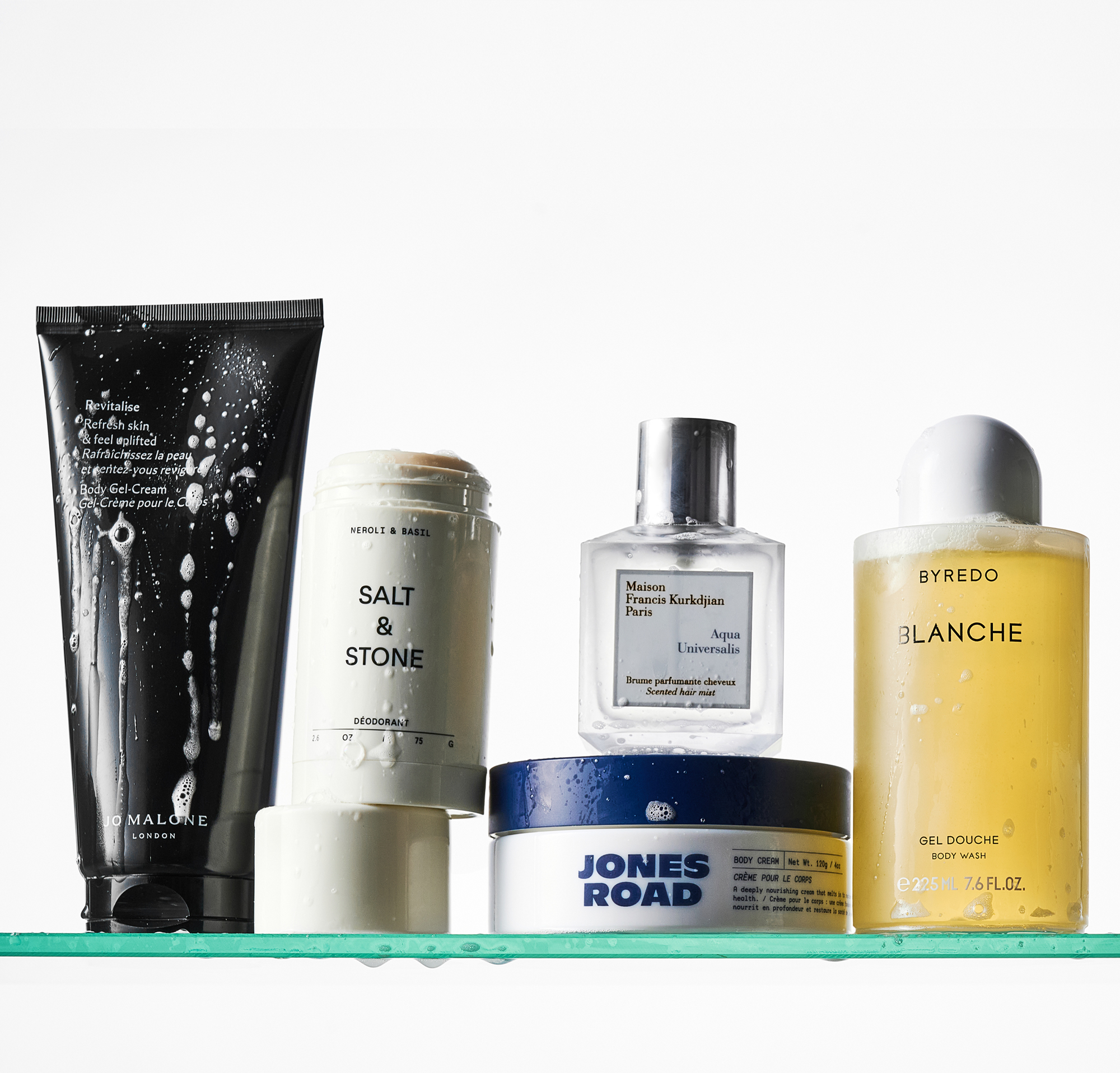 A Jo Malone body lotion, Salt &amp;amp; Stone deodorant, Jones Road body cream, MFK hair mist and Byredo body wash on a glass shower shelf