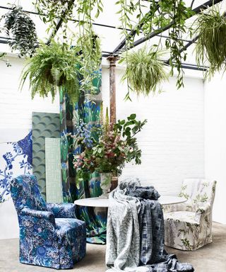 Hanging baskets in a white patio garden with green and blue patterned textiles draped around a table and chairs.
