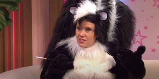 Kate McKinnon dressed as skunk Pepe Le Pew with her face exposed, smoking a cigarette and looking amused.